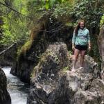A picture of Caitlyn Grubbs standing on a boulder next to a stream in the woods