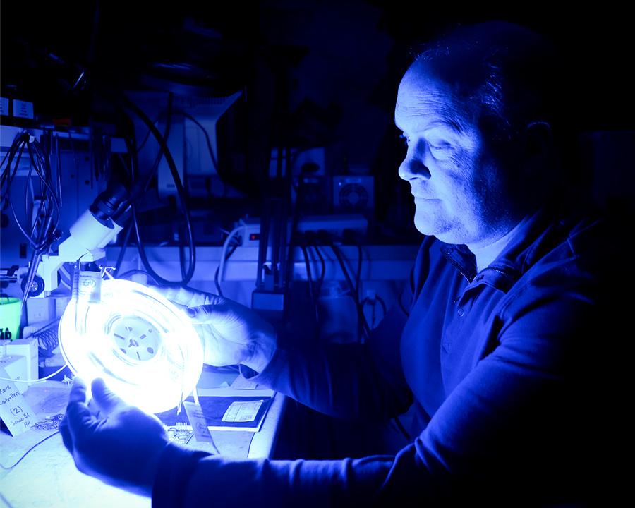 electronics staff examines project. Blue glow from light illuminates a dark room.
