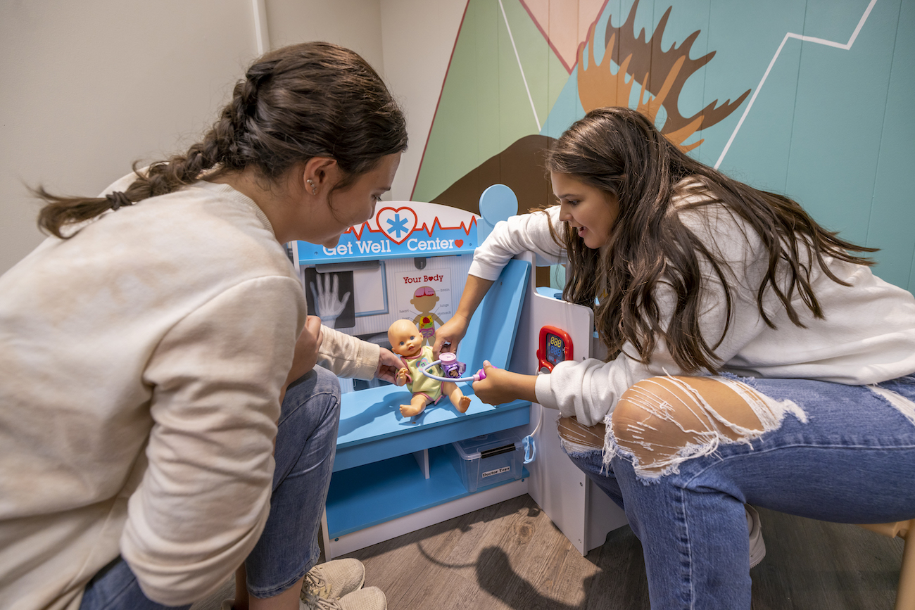 Two students in the play lab. They use a child's nurse station to check the heartbeat of a plastic baby doll.