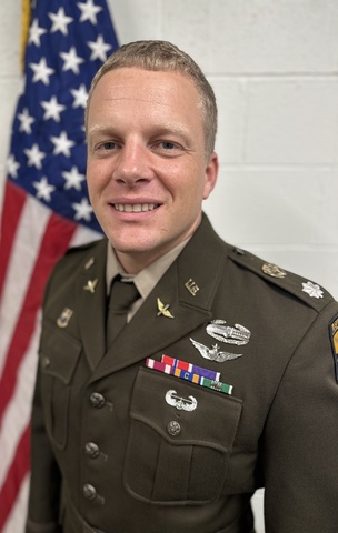 A portrait of Todd Kuebler in a military uniform standing in front of the American flag