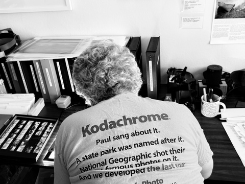 A black and white photo of Johnny Brian Pasierb examining photos with his back facing the camera. His tee shirt reads: "Kodachrome. Paul sang about it. A state park was named after it. National Geographic shot their most famous photos on it. And we developed the last roll. Dwayne's Photo: we made history on December 30, 2010.