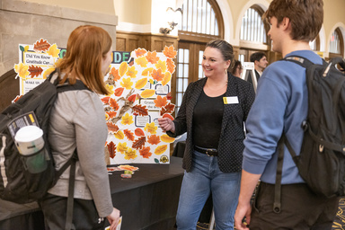 Emily standing with two students at a table in front of a poster board with colorful leaves.