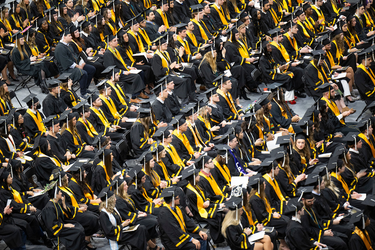 A wideshot of the students sitting in rows, facing the stage, wearing their caps and gowns