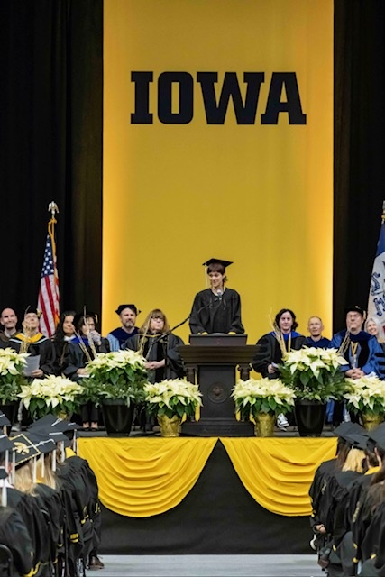 Student commencement speaker stands on stage at commencement in front of a bold Iowa banner