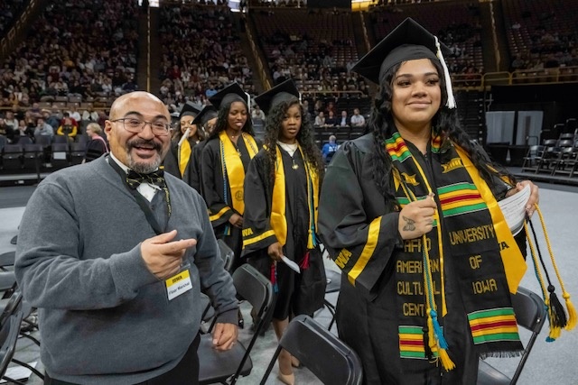 A man smiles, standing next to a female graduate adorned in a custom embroidered stole