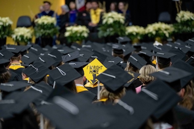 A vibrant yellow mortarboard amongst a sea of black caps. It reads "I'm Right Here."