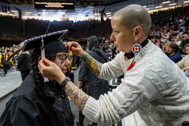 A family member adjusts a beaded adornment on a graduate’s mortarboard during a University of Iowa commencement ceremony.