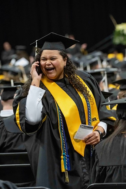 student in black and gold commencement attire wears an enthusiastic grin, standing in commencement hall on her cellphone