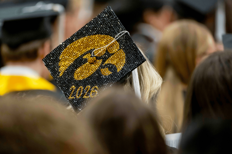 A graduate decorated their black commencement cap with a sparkling gold Hawkeye logo and the year “2026.” The cap stands out in a crowded ceremony scene filled with graduates and attendees seated nearby.