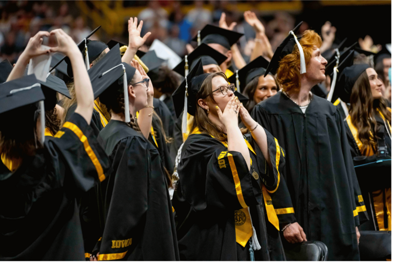 Graduates in black caps and gowns react during a commencement ceremony, with several waving or making heart shapes with their hands toward the crowd. One graduate in the center blows a kiss while classmates around them smile and look into the audience.