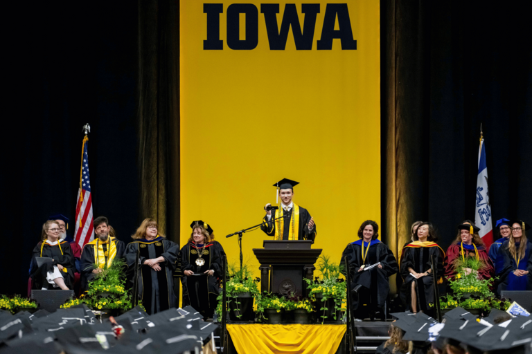 A student speaker stands at a podium during a commencement ceremony beneath a large gold banner reading “IOWA.” Faculty and university leaders in academic regalia sit on stage behind the speaker holds a camera back at the crowd, while graduates in caps and gowns fill the foreground.
