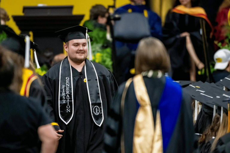 A graduate wearing a black “First Generation” stole walks across the commencement floor during the ceremony. Faculty members and fellow graduates in academic regalia surround the stage area in the background.