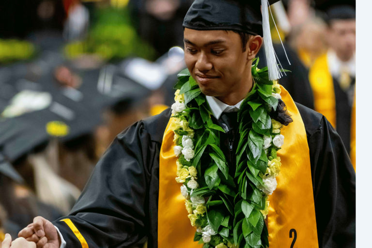 A graduate wearing a black cap and gown, gold stole, and green floral lei reaches out to shake someone’s hand during the commencement ceremony. Other graduates in caps and gowns are visible in the softly blurred background.