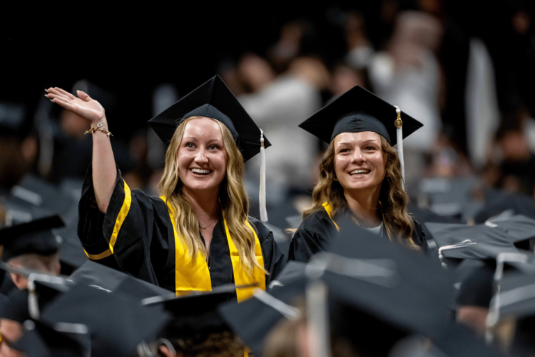 Two graduates in black caps and gowns smile from the audience during a commencement ceremony. One graduate waves toward the crowd while both look out into the arena, surrounded by fellow graduates seated nearby.