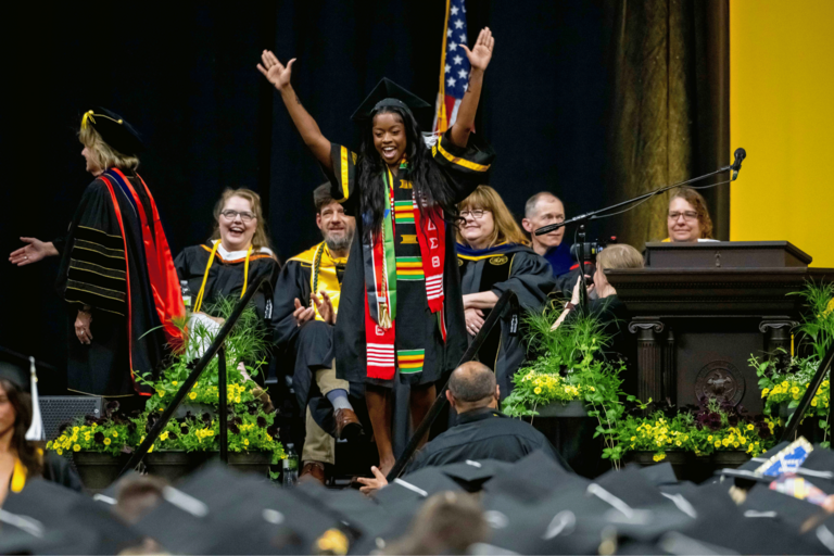 A graduate wearing colorful stoles raises both arms in celebration while walking across the commencement stage. Faculty members and university leaders seated behind the stage smile and applaud as graduates in caps and gowns fill the foreground.