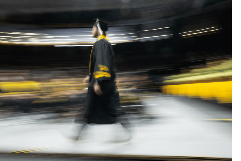 A graduate in a black cap and gown walks across the commencement floor as the surrounding arena blurs with motion. The image captures the movement and pace of the ceremony, with streaks of light and rows of seated attendees in the background.