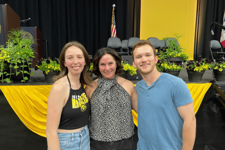 CLAS Dean Sara Sanders poses with the student commencement speakers from the morning and afternoon ceremonies in front of the graduation stage during rehearsal. The three smile for a photo beside black-and-gold Commencement decorations and rows of flowers after the ceremony.