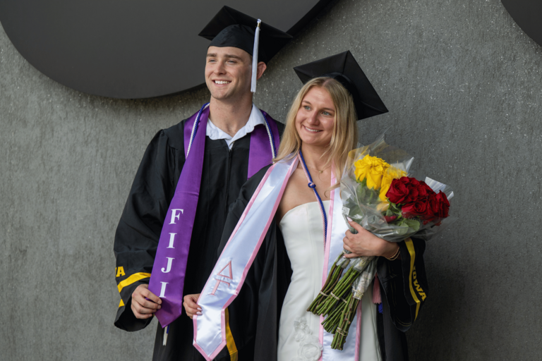 Two graduates in black caps and gowns pose together in front of a textured gray wall. One graduate wears a purple stole with “FIJI” lettering, while the other wears a white stole with pink trim and holds bouquets of yellow and red roses. Both are smiling and looking slightly off camera.