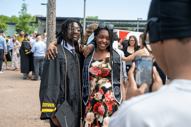 A graduate in a black cap and gown poses with a smiling companion in a floral dress and graduation stole during an outdoor Commencement gathering. Another person in the foreground holds up a phone to take their photo, while groups of graduates, families, and friends gather in the background.