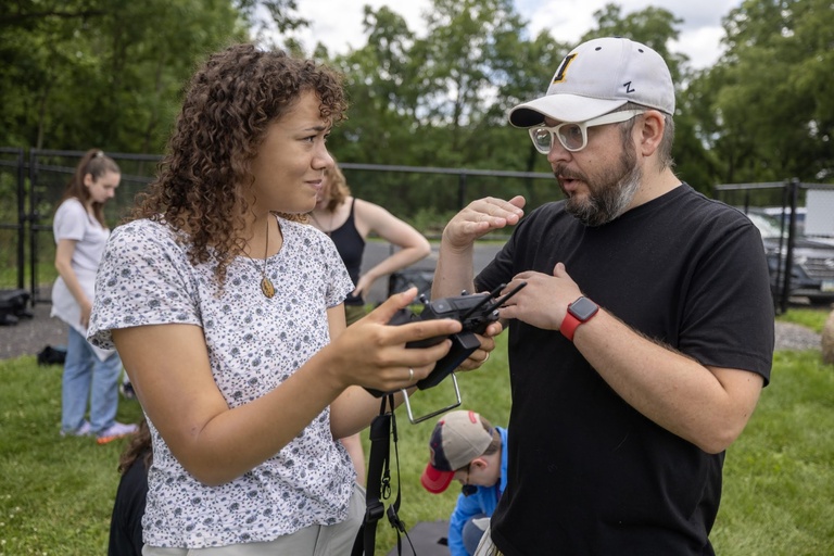 Adam Skibbe explains how to operate a drone controller to a student during an outdoor field activity.