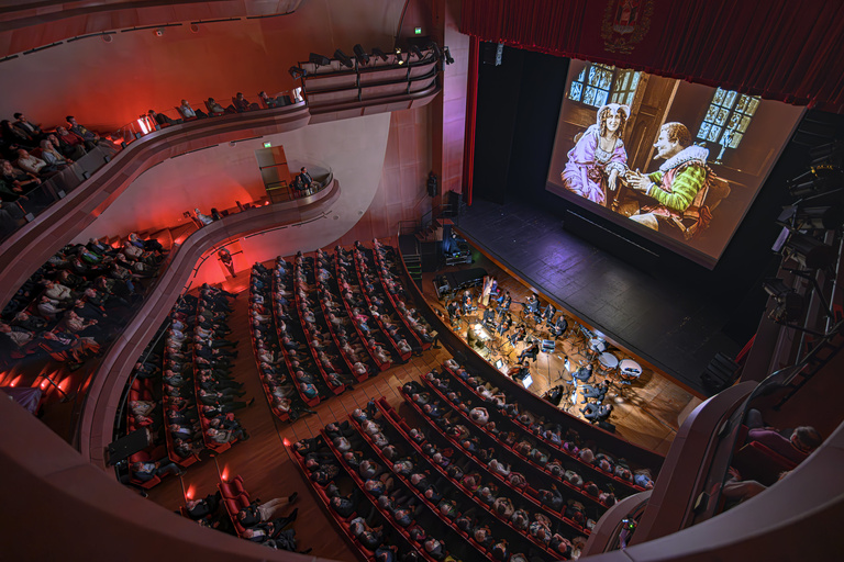 Overhead image of a theater, which shows the audience, orchestra in the pit, and a film screen with a movie playing.