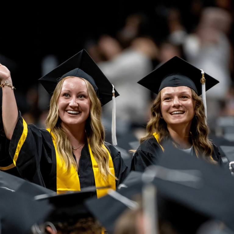Two graduates in black caps and gowns smile from the audience during a commencement ceremony. One graduate waves toward the crowd while both look out into the arena, surrounded by fellow graduates seated nearby.