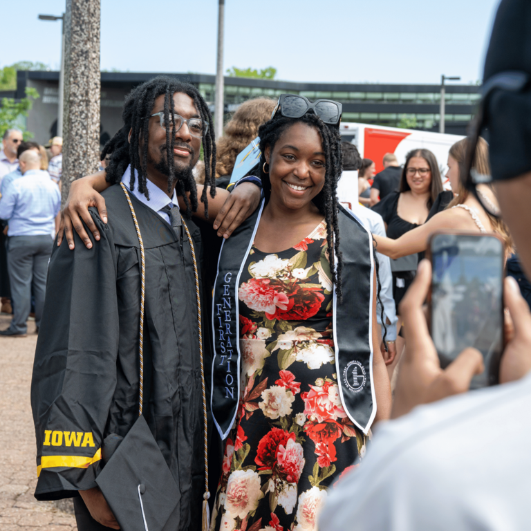 A graduate in a black cap and gown poses with a smiling companion in a floral dress and graduation stole during an outdoor Commencement gathering. Another person in the foreground holds up a phone to take their photo, while groups of graduates, families, and friends gather in the background.