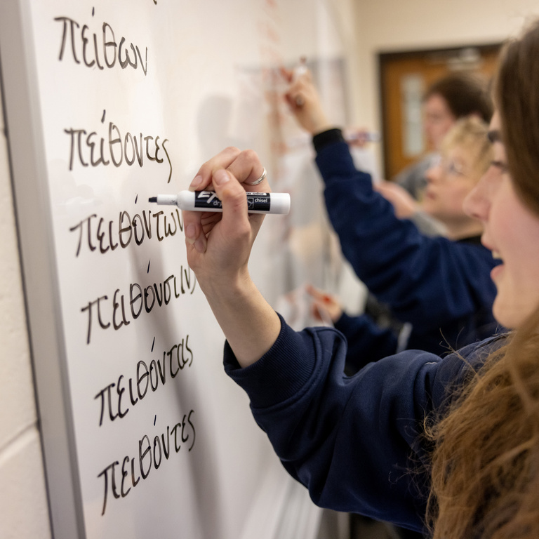 A student writes forms of an Ancient Greek verb participle in marker on a whiteboard, working through case and number endings 