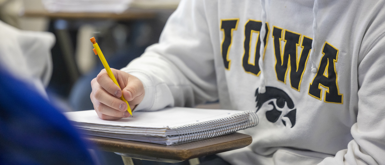 Student sitting at a desk in a classroom with a pencil and notebook wearing a University of Iowa sweatshirt