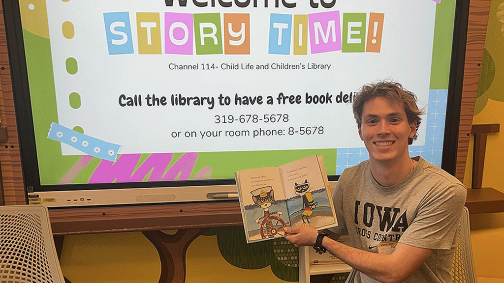 Photograph of Will Ryan holding an open children's book while seated in front of a drop down screen reading "Story Time"