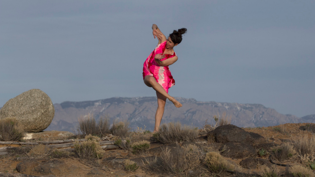 Stephanie Miracle dances barefoot on a rocky hillside, with mountains and open sky in the background.
