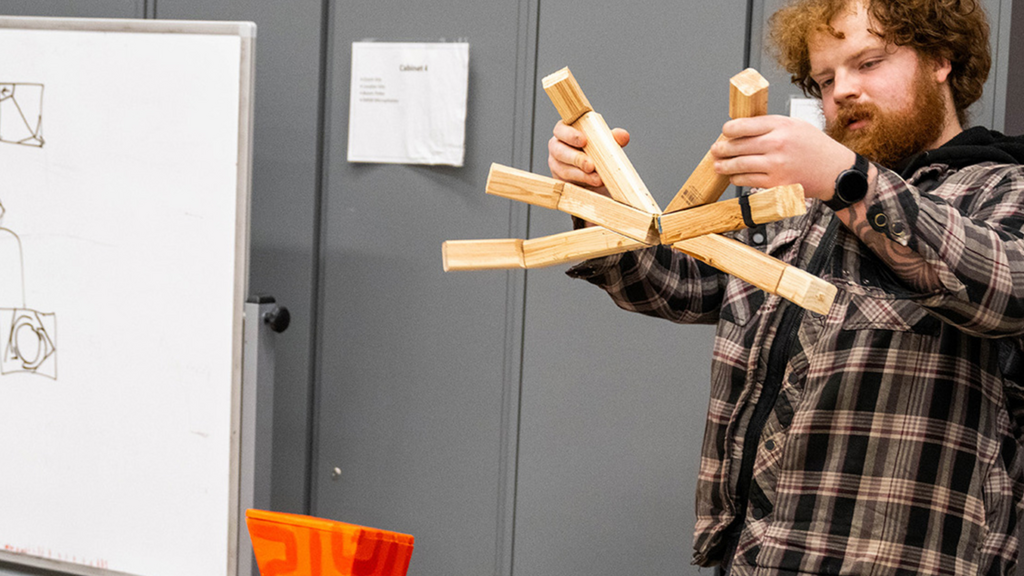 A student demonstrates a wooden articulated prototype with several hinged arms while standing in a workshop. A whiteboard beside them displays a hand-drawn sketch of the machine's design, and an orange geometric prototype sits on a chair in the foreground, illustrating the progression from concept sketch to physical model