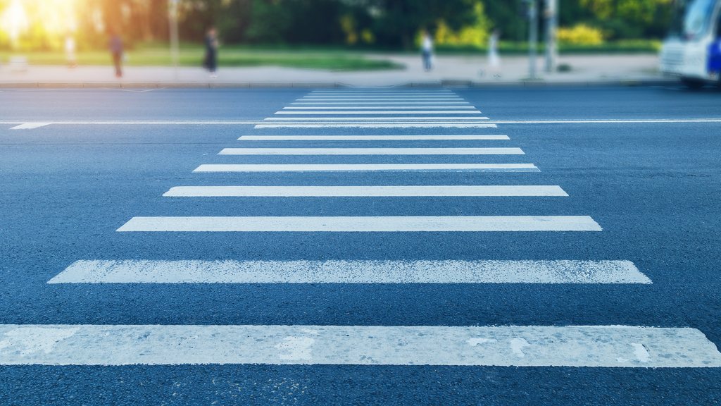 A white-striped pedestrian crosswalk stretches across a wide asphalt road toward a tree-lined sidewalk. The background is softly blurred, with a few pedestrians waiting near the curb and part of a bus visible on the right, while warm sunlight shines from the upper left.