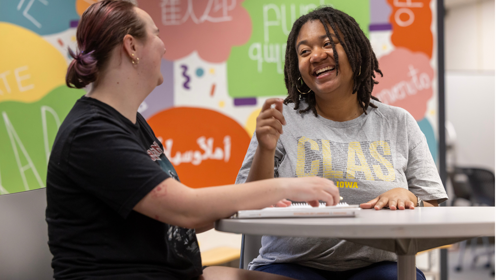 two students sit together in front of a green, yellow, blue, orange, and red backdrop. The students are in coversation and laughing at their desks.